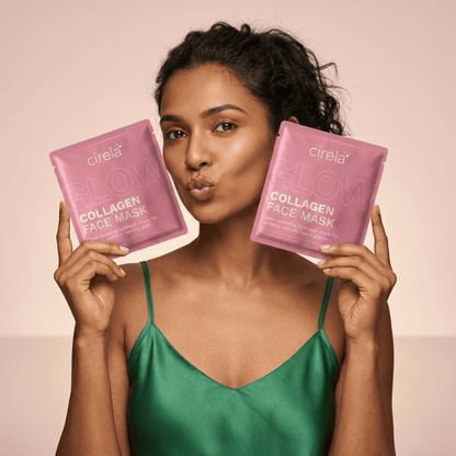 A woman in a green satin top holds up two pink Glow Collagen Hydrogel Face Mask packets from Cirela Beauty, smiling and making a kissy face against a neutral background—ready for radiant, glowing skin.