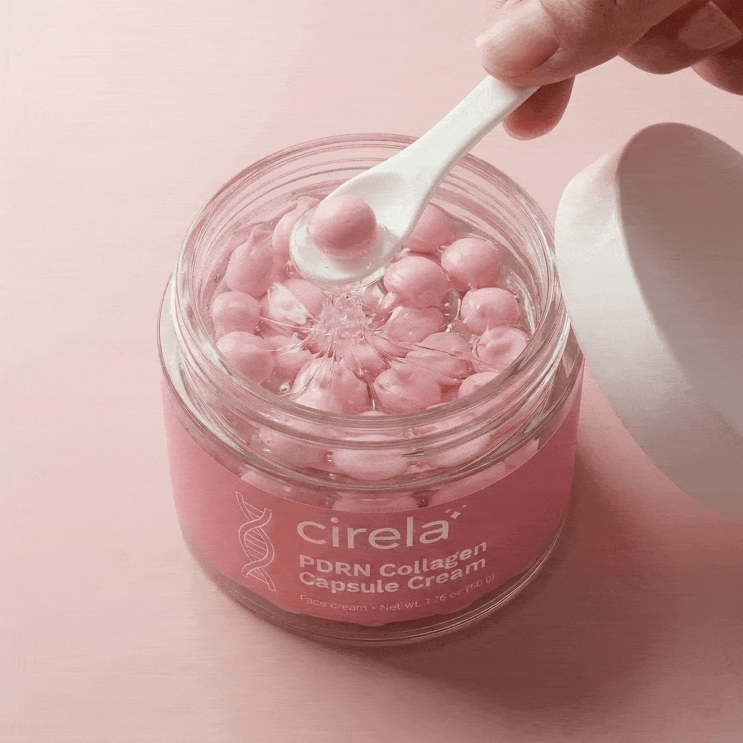 A hand holds a small white spoon scooping a pink capsule from a Cirela Beauty PDRN Collagen Capsule Cream jar, filled with shiny pink capsules for instant glow and even skin tone, against a pink background.