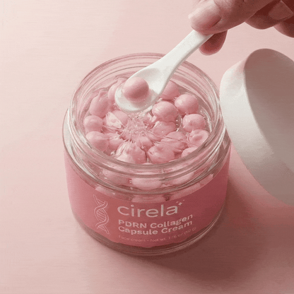A hand holds a small white spoon scooping a pink capsule from a Cirela Beauty PDRN Collagen Capsule Cream jar, filled with shiny pink capsules for instant glow and even skin tone, against a pink background.