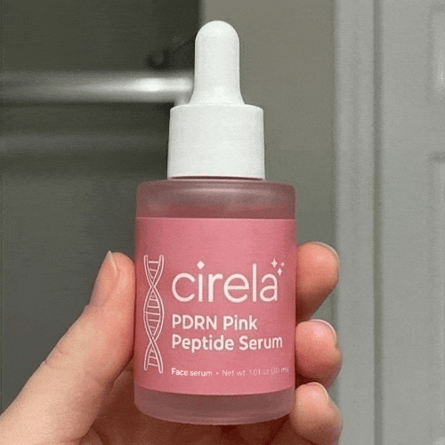 A hand holds a small pink bottle of Cirela Beauty's PDRN Pink Peptide Serum with a white dropper cap, offering lightweight hydration. The background is blurred, suggesting a bathroom setting.