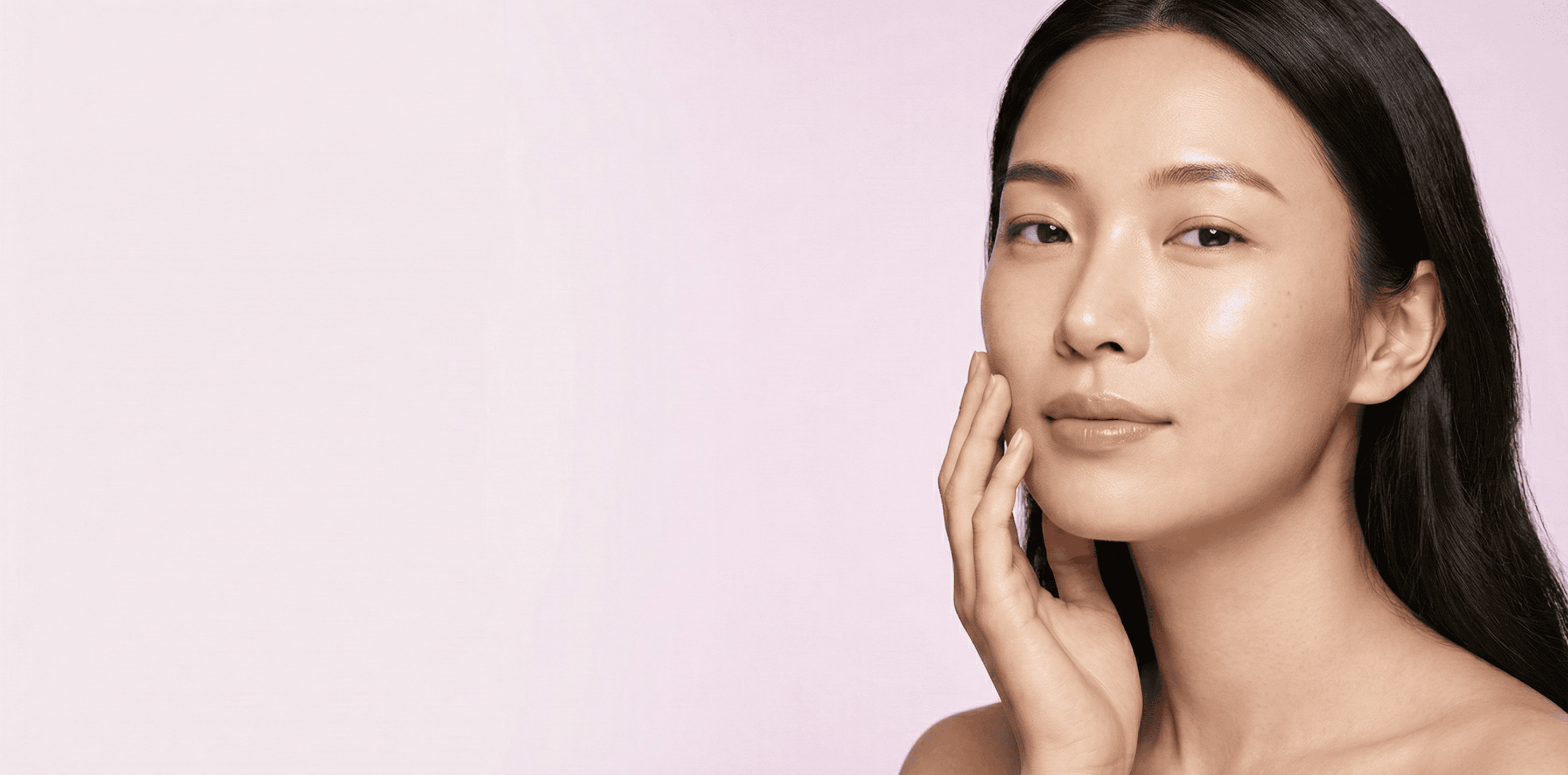 A woman with long, straight black hair touches her face gently, showing smooth, radiant skin. She looks confidently at the camera against a soft pink background.