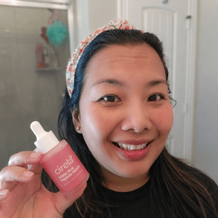 A smiling person with long dark hair and a floral headband holds up a dropper bottle labeled Cirela Beauty PDRN Pink Peptide Serum in a bathroom setting.