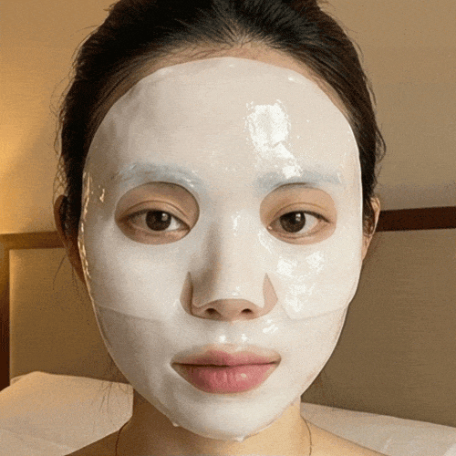 A woman with straight dark hair wears the Glow Collagen Hydrogel Face Mask by Cirela Beauty, gazing at the camera in a well-lit room and showcasing her radiant, pore-smooth skin after use.